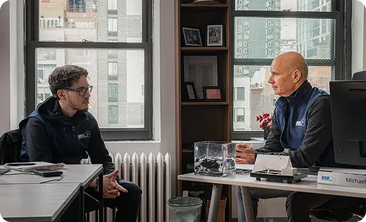 Two men sit at a desk in an office discussing a small terrarium-like container between them