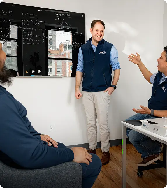 Man in a blue vest stands smiling while presenting to two colleagues in an office with a blackboard