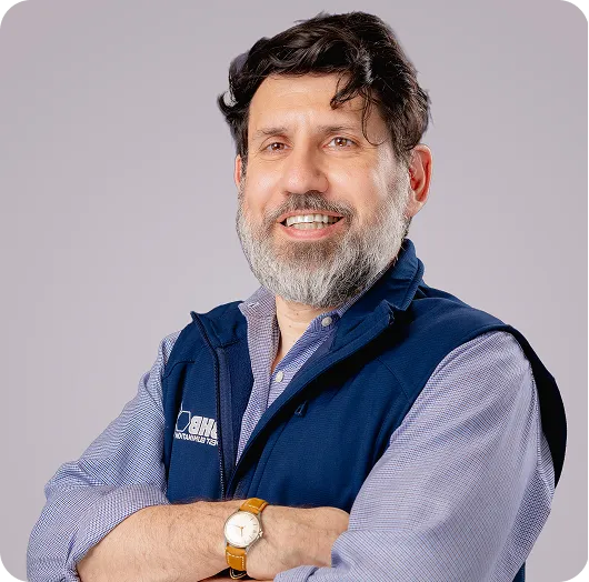 Smiling man with gray beard, arms crossed, wearing a navy vest over a blue shirt