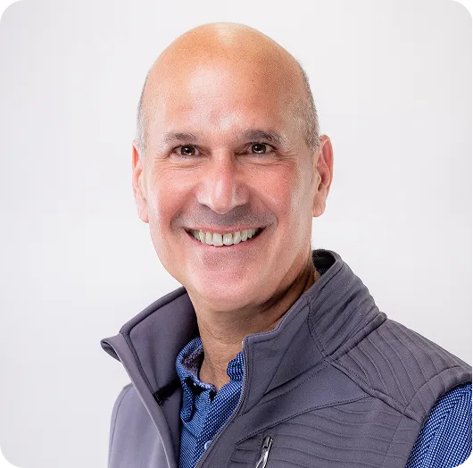 Smiling bald man in a gray vest over a blue plaid shirt, posed for a professional headshot