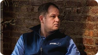 Man in a navy vest over a blue shirt sitting indoors in front of a brick wall